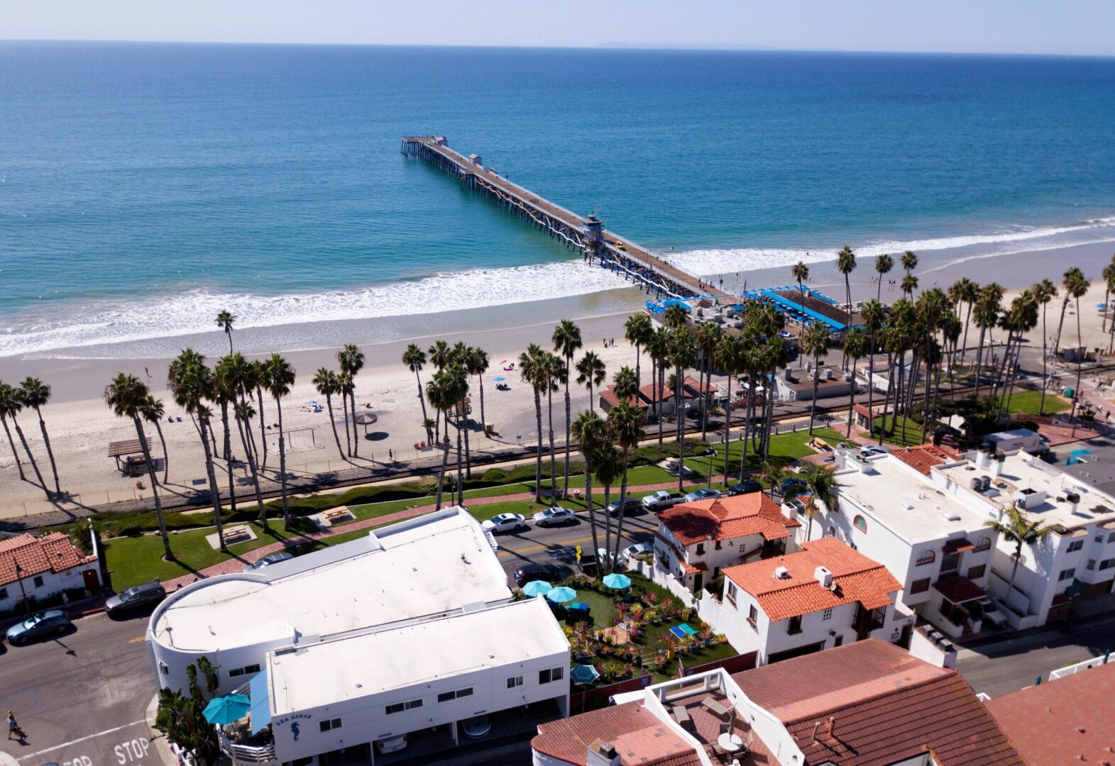 A view of the ocean from above shows palm trees, buildings and pier.